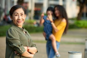 Waist-up portrait of cheerful mature woman posing for photography with arms crossed, blurred silhouettes of curly woman holding little child on arms and pointing at something with index finger